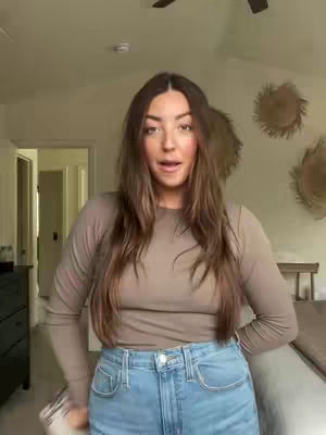 A woman with long brown hair stands in a bedroom facing the camera, talking. She wears a long-sleeved beige top and blue jeans. The background includes a bed, wall decor, and an open doorway.
