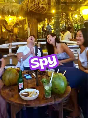 A group of four people are sitting around a wooden table outdoors in a tropical setting. There are two large green coconuts on the table along with drinks and food. The text overlay says "Spy" with a purple and white color scheme, indicating a social deduction game. Two people are laughing.