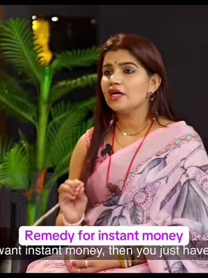 A woman in a pink saree is speaking, with text overlayed at the bottom that reads: 'Remedy for instant money'.  Behind her is a green plant. The background appears to be a dark room.