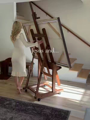 A person stands in front of a wooden easel with a large framed canvas. The person is seen from the back and is wearing a long white dress. They are in a house with light-colored walls, the composition is well-lit and creates a sense of a private moment or artistic process and there are stairs leading upstairs.