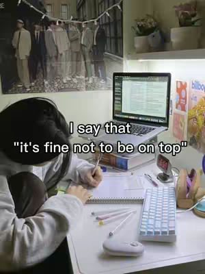A person is seated at a desk, writing in a notebook with a pen. The desk is cluttered with items like a keyboard, pens, a mouse, and a small desktop clock and a laptop. A poster is visible in the background, and the overall lighting suggests a well-lit room.