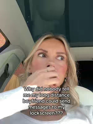 A selfie-style shot taken from inside a vehicle. A blonde woman is in the foreground, appearing surprised or shocked, with her right hand covering her mouth. The background shows a blurry view through the car's windshield and the interior of the car. Text overlayed on the image asks why nobody told the creator that her long-distance boyfriend could send messages to her lockscreen.