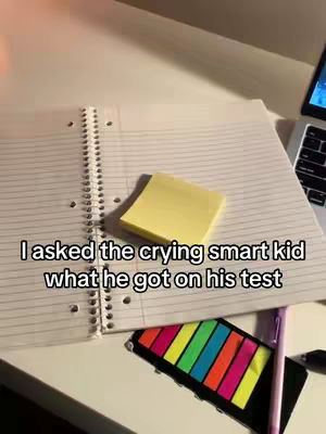A top-down shot of a white desk with a laptop, notebook, sticky notes, colored flags, pens, and a laptop. Text overlayed on the image reads: "I asked the crying smart kid what he got on his test".
