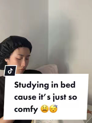 The cover image shows a person, likely female, lying in bed with a black head covering. A white rectangle with black text overlay is placed, the text reads 'Studying in bed cause it's just so comfy.' There are emojis showing a tired face and a sleeping face.