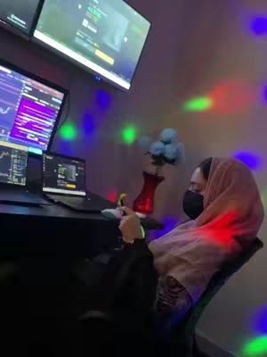 The image shows a woman, wearing a hijab and a face mask, seated at a desk with a laptop and multiple monitors displaying what appears to be financial data. The scene is dimly lit with colored lights creating a party atmosphere.