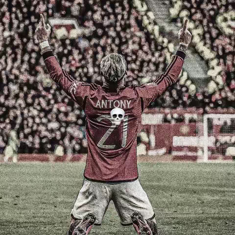A soccer player in a red jersey with the name "ANTONY" and number "21" on the back, standing on a grassy field. The player has his back to the camera, arms raised in celebration with both hands making the peace sign or the middle finger. The backdrop shows a crowd of people blurred in a stadium.