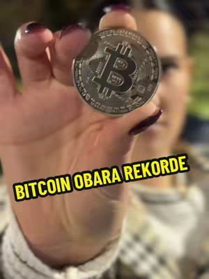 A close-up shot showcases a hand holding up a silver Bitcoin coin with the Bitcoin logo clearly visible. Dark red nail polish adorns the fingers, and the background is slightly blurred, suggesting a focus on the coin and creator.