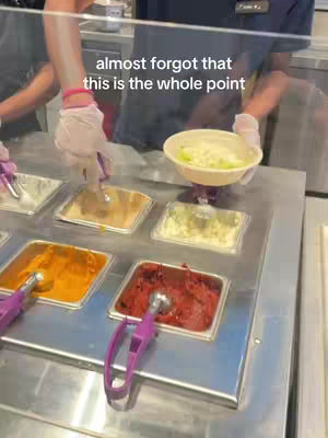 A person wearing a uniform in a food service area is shown from the waist up. They are behind a display case filled with ingredients. The person holds a bowl with ingredients and two gloved hands. The background has food containers and other staff members.
