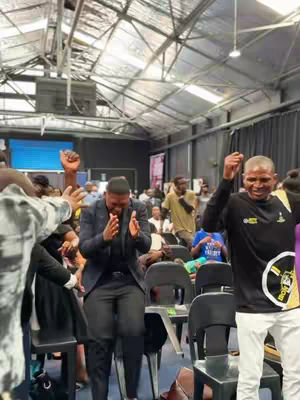 A photo from inside a large, open church or event space. The ceiling is high, with exposed metal beams and a translucent roof. People are gathered, some seated and many standing, raising their hands in the air. Several individuals, likely church leaders or speakers, are in the foreground.