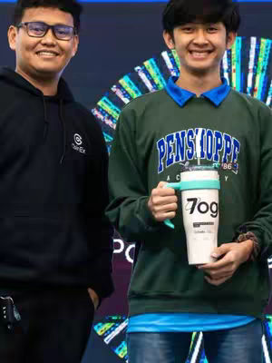 Two young men standing in front of a blurry background, one on the left wearing glasses and a black hoodie, and the other on the right with a green sweater holding a tumbler. They are both smiling and looking towards the camera.