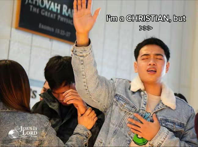 A selfie-style shot featuring a young Asian man in a denim jacket, with closed eyes and an expression of religious fervor, his right arm raised. Behind him, two other people are partially visible, also with folded hands, suggesting a church or religious setting. The image includes a text overlay and a logo.