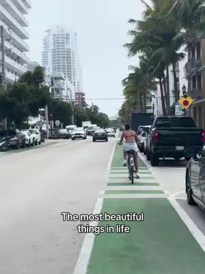 A woman rides a bicycle down a city street with tall buildings on either side. A green bike lane is painted on the right side of the road. The sky is overcast, and the overall impression is calm.