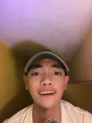 A young man with a green cap and white shirt, looking at the camera and talking. The background is a two-tone tan and purple. A microphone is in front of the man.