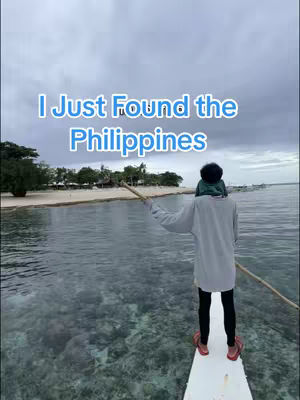 The image shows a person standing on a small platform extending from a boat, holding a wooden pole, with their back to the viewer. They are looking out at the ocean. The water is clear, revealing the sea floor. In the background, there's a sandy beach with some trees and a cloudy sky overhead. Overlay text in a playful font spells, "I Just Found the Philippines."