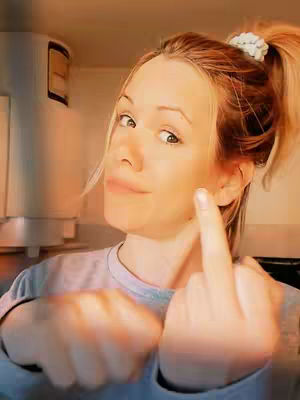 A selfie-style shot featuring a woman with blonde hair pulled back in a high ponytail with a white scrunchie. She is wearing a gray shirt and pointing a finger directly at the camera, with the middle finger up in a gesture. The background is out of focus.