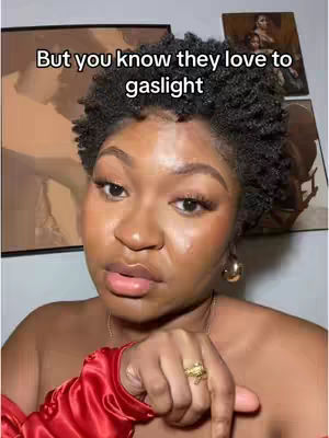 A selfie-style cover image features a Black woman with short, dark, curly hair. She has full lips, prominent cheekbones, and is wearing makeup. She looks directly at the viewer with a thoughtful expression. A text overlay at the top says, "But you know they love to gaslight." Her attire includes a red satin top, and she wears gold hoop earrings and a gold ring with a unique design. The background includes framed pictures on the wall.