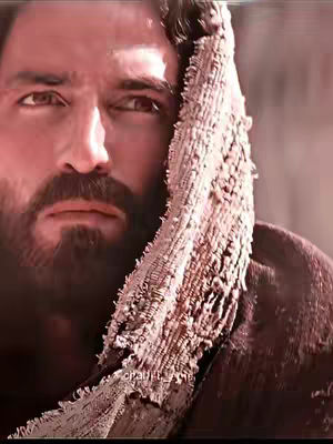 A close-up shot of a man with a beard, dark hair, and brown eyes. He appears to be shrouded in a cloth, with sunlight casting shadows on his face. The overall aesthetic is serious and dramatic.