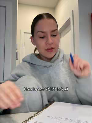 A woman with brown hair pulled back is sitting at a desk with a notebook, holding a blue pen. The background is a blurred, neutral space; the image focuses on the woman and the notebook.