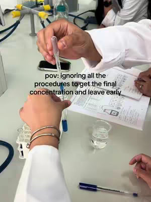 The image shows a first-person perspective of a laboratory setting. In the foreground, a person's hands are visible; one hand is holding a pipette, dispensing blue liquid into a test tube, while the other arm has bracelets. Papers containing lab instructions and a beaker with clear liquid are on the table in the background. The setting is a mix of lab equipment.