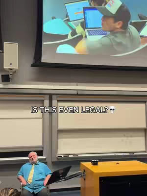 A classroom setting with a projector screen displaying a man using a laptop. In the foreground, there is a man sitting in a chair with a laptop on a stand in front of him.