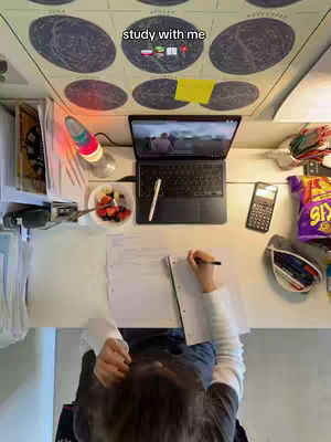 An overhead shot of a desk setup with a person studying. A laptop is open, showing a video, next to a pen and notepad. There's a half-eaten bowl of fruit and a calculator. The background includes a map and a decorative lamp.
