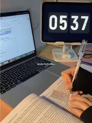The image shows a desk setup with a laptop open in the foreground, displaying what appears to be a document with lines of text. A book lies open beneath the laptop, its pages filled with text, and a pen rests on the book. To the right, a tablet displays the time 05:37, and a soft light illuminates the scene, suggesting a study or work environment.