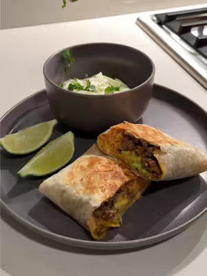 A close-up shot of a plate with a partially cut burrito, two lime wedges, and a small bowl of dip. The burrito is cut in half, revealing a filling of what appears to be meat, avocado, and cheese. The bowl contains a white dip, with flecks of green herbs. The lime wedges are bright green. The plate is a muted gray color, and the background fades out of focus.