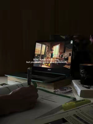 A dimly lit desk with a laptop showing a video of a well-lit room on screen. A hand is visible working on a notebook while a pen and highlighter rest on the desk.