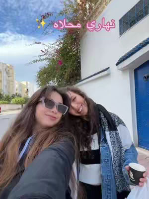 Two young women taking a selfie outdoors. They are positioned close together, with one slightly in front, both looking at the camera and smiling. The background includes a building with a blue door, a wall, and some greenery, under a partly cloudy sky.