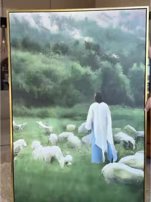 A framed artwork featuring a figure in a white robe, positioned on a green field surrounded by sheep. The background shows a blurry, green, and slightly hazy landscape suggesting mountains or a forested area. The artwork has a gold frame. The main figure is seen from the back.