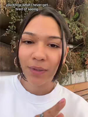 A close-up shot of a woman's face. She is looking towards the camera and speaking. The background shows a wall covered in plants. The woman is wearing a white sweatshirt and hoop earrings. She's also wearing a ring on her finger and has pink nail polish on one hand. The hook text is overlaid on the top of the image.