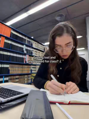 A young woman is seated at a desk in a library setting, writing in a notebook. There is a laptop and a dark object on the desk in the foreground. Bookshelves fill the background.