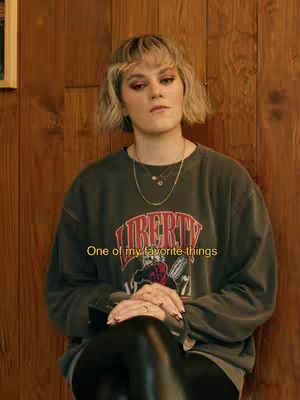 The image features a woman seated indoors, with a wooden paneled wall forming the backdrop. She wears a gray sweatshirt with the word "LIBERTY" in bold letters on the front, combined with black leather-like pants. She has blonde hair and clear makeup. A text overlay appears on the image, adding a quote. The overall composition presents a casual, contemporary style.