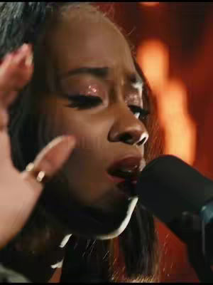 A close-up shot of a woman singing into a microphone. Her face is partially visible, with a slight smile, and her hand, adorned with rings, is raised in front of her. The backdrop suggests a performance setting with blurred lights.