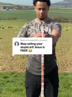A man stands in a field with a wooden rod in front of him. A text bubble originating from a comment covers his torso. The background shows fields, trees, and a glimpse of a distant building under a bright blue sky.