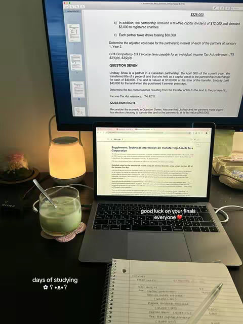 The image shows a desk setup with a focus on studying for finals, including a laptop displaying study notes, a notepad with handwritten notes, and a glass of green drink. The setup is well-lit, with an overhead light fixture.