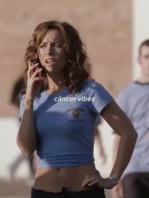 A woman with curly brown hair talks on a pink phone in a sunny outdoor setting. She wears a blue shirt and has her hand on her hip. The text 'câncer vibes' is overlaid at the bottom.