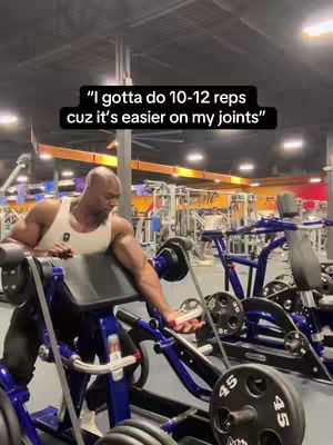A muscular man with a shaved head is working out in a gym. He's using a machine that appears to be a seated preacher curl setup. Above him, a text overlay reads, "'I gotta do 10-12 reps cuz it's easier on my joints'". The gym is well-lit, with various exercise machines and equipment visible in the background.