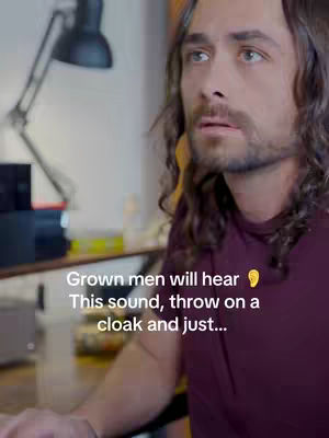 A medium shot shows a man with long, wavy brown hair gazing off-camera with a somewhat wistful expression. He is wearing a burgundy t-shirt in a dimly lit setting, likely indoors. The background is blurred, but visible are elements suggestive of a desk or workspace, including a desk lamp. Text overlayed on the image reads, 'Grown men will hear 👂 This sound, throw on a cloak and just...'
