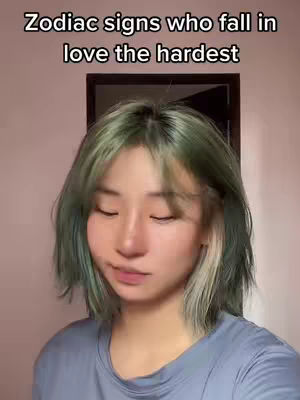 A woman with dyed green hair looks down with closed eyes in front of a tan wall and a dark brown picture frame, appearing to be in a blue shirt.