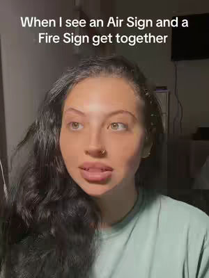 A woman with wavy dark hair and a light green shirt looks up and to the left with a thoughtful expression. Above her, text overlays read, “When I see an Air Sign and a Fire Sign get together”. The background is dark and out of focus, suggesting an indoor setting.