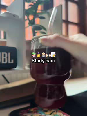 The image shows a close-up of a glass and a hand on the top. The glass is full of a red liquid. Behind the glass is a JBL speaker, a plant, and a window with some light coming in from the outside. Text overlayed on the image says 'Study hard'.