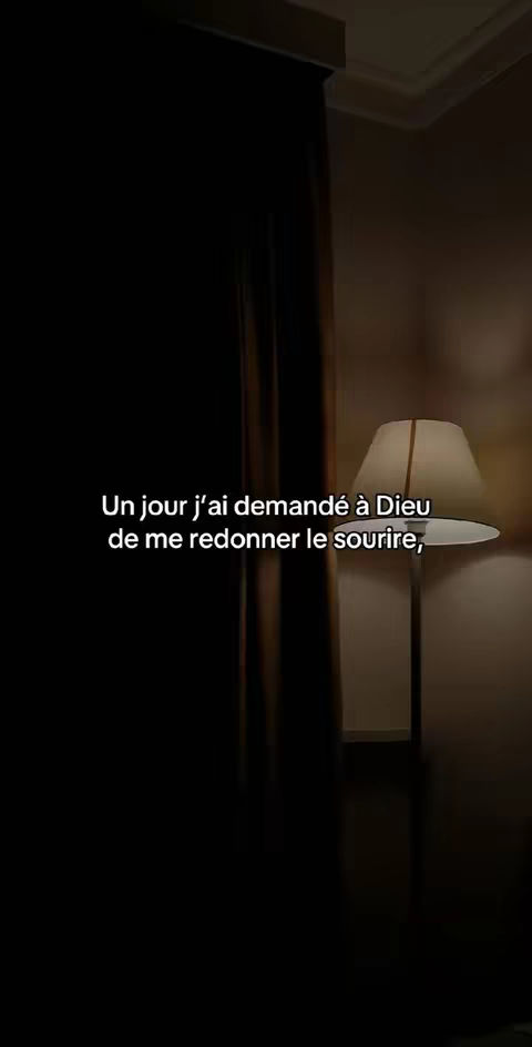 The image shows a dimly lit room with a dark curtain on the left side. In the background, a lit table lamp is visible, casting a warm glow. The text overlayed on the image reads "Un jour j'ai demandé à Dieu de me redonner le sourire,"
