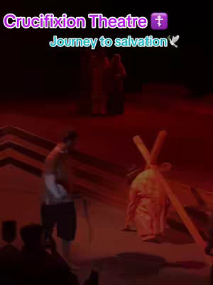 A stage performance depicting a crucifixion scene. The image is dominated by a deep red color scheme, suggestive of a dramatic and somber mood. In the foreground, a figure, possibly representing Jesus, carries a cross, while other figures are visible in the background on a stage. Text overlays at the top indicate the video is about a 'Crucifixion Theatre' and a 'Journey to salvation.' The composition is simple, focusing on the central act of the performance.
