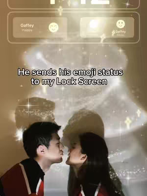 The image shows a couple about to kiss, silhouetted against a light effect that resembles a cloud. At the top of the image, there is a phone interface showing two emoji status icons. The overlay text reads, "He sends his emoji status to my Lock Screen". The overall color tones are warm and golden.