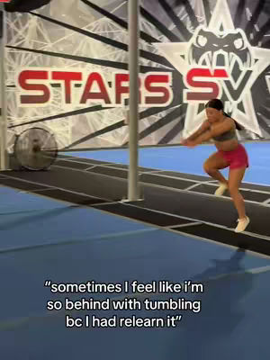 A young woman is mid-jump, captured in motion inside a cheerleading gym. She wears a pink sports bra and shorts, with her arms extended forward. The setting includes a blue and black floor, a wall featuring the text 'STARS SV' in bold red and white letters, and a large fan. Text overlays the bottom of the image.