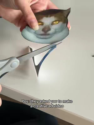 A hand holds a cat face cutout above a piece of paper while cutting an arrow shape, a pair of scissors are open and ready to cut the paper. The scene is against a white surface.