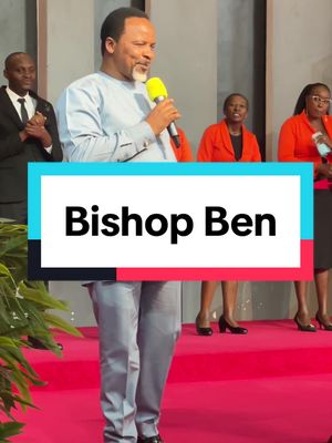 A man in a light blue shirt is speaking into a yellow microphone, with a backdrop of a stage and several people behind him. Below him is a text overlay that reads "Bishop Ben". The scene is bathed in warm lighting, indicative of an indoor event.