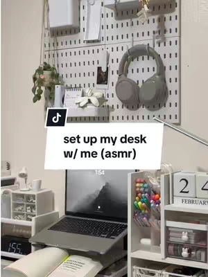The image shows a well-organized desk setup with a focus on aesthetics and productivity. A laptop sits on a stand, and there's a pegboard on the wall with headphones and other items. The color palette is predominantly white and light-toned, with pops of color from pens and other desk accessories. The composition is clean and inviting, suggesting a workspace designed for study or creative work.