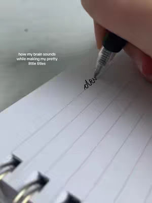 The cover image shows a close-up view of a hand writing in cursive with a black pen on a lined notebook page. The pen is mid-stroke, forming the beginning of a word. The image has a simple, clean aesthetic with a neutral color palette dominated by white and black. The text overlay is in the upper left corner against a blurry, white background.